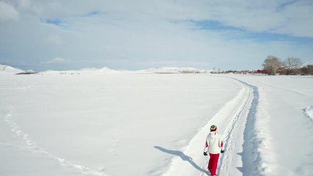 Aerial view; drone flight, camera moves behind girl going forward on the frozen lake; young woman walking among snow field in red pants and white sport jacket; calm, happy peaceful mood; beauty around
