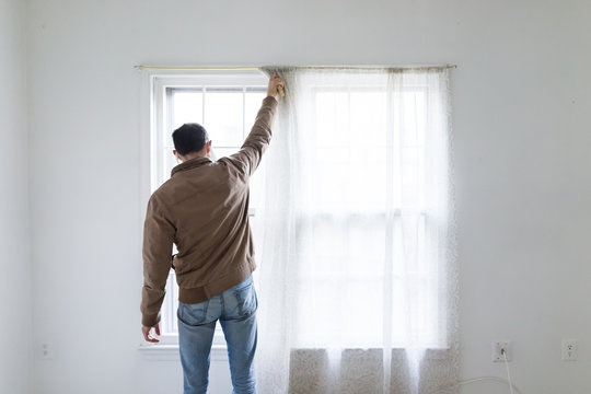 Young man standing in front of bright window in room adjusting blinds curtains, white painted walls during remodeling removing cleaning inspection