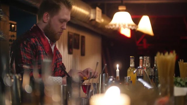 Young professional bartender wearing red apron putting a lemon into a glass with red liquor