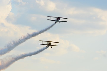 Two stunt biplanes flying one above the other on a cloudy blue sky leaving white smoke trail