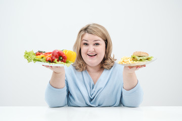 Young lush fat woman in casual blue clothes on a white background holding a vegetable salad and a plate of fast food, hamburger and fries. Diet and proper nutrition.