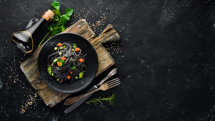 Black pasta with vegetables on a black plate. Top view. On a black background. Free copy space.