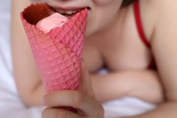 Sexy woman eating ice cream waffle cone in the bed, sweet tooth. Girl in lingerie enjoys the strawberry cream, tasty dessert