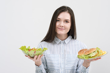 Slender brunette girl on a white background chooses between a plate of fast food and hamburgers and healthy food salad.