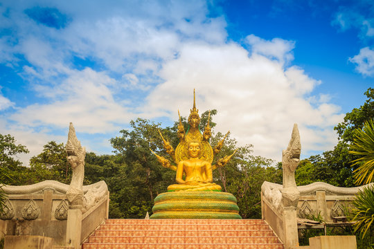 Beautiful golden Buddha statue with seven Phaya Naga heads under white clouds and blue sky background. Outdoor golden seated Buddha image protected by 7 heads Naga spreads cover on top in cloudy days.