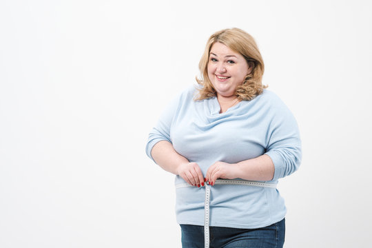 A fat woman measures her waist with a measuring tape in casual clothing on a white background.