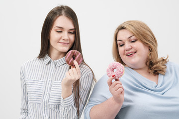 Two girls, thin and fat on a white background, are holding pink glazed donuts in their hands.