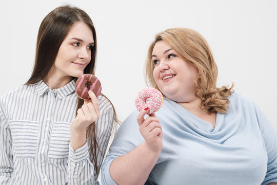 Two girls, thin and fat on a white background, are holding pink glazed donuts in their hands.