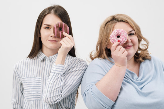 Two girls, thin and fat on a white background, are holding pink glazed donuts in their hands.