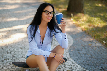 Portrait of a beautiful girl in a white shirt and shorts holding a cup of takeaway coffee and looking at a camera while sitting on a skateboard in the park on a summer morning.