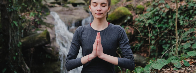 Naklejka na ściany i meble Young slim woman practicing yoga outdoors in moss forest on background of waterfall. Unity with nature concept. Girl standing with prayer hands