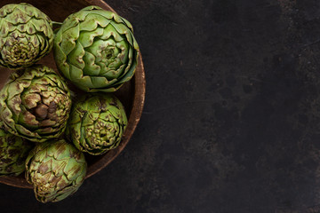 Close up photo of fresh artichoke in the old wooden bowl. Top view on dark background - obrazy, fototapety, plakaty