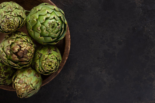 Close up photo of fresh artichoke in the old wooden bowl. Top view on dark background
