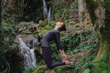 Naklejka na ściany i meble Young slim woman practicing yoga outdoors in moss forest on background of waterfall. Unity with nature concept. Girl in camel position