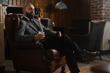 Portrait of serious bearded man with pipe holding glass of whiskey wearing suit and sitting on a big arm chair