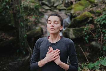 Young woman practicing breathing yoga pranayama outdoors in moss forest on background of waterfall. Unity with nature concept.