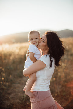 Motherhood - young mother hugging her happy baby in fields at sunset. Woman kissing her smiling barefoot baby boy outside in the nature on summer day.