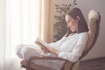 Candid portrait of a woman resting in a chair by the window, reading a book. Wellness, education, self-development