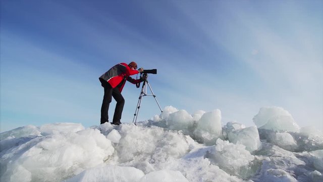 Sliding view from below of photographer with camera on ice blocks taking photos or video of frozen lake. Blue sky is on the background