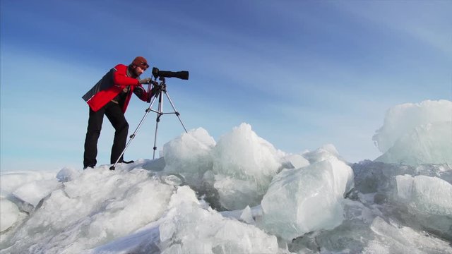 Sliding view from below of photographer with camera on huge ice blocks taking photos or video of frozen lake. Blue sky is on the background
