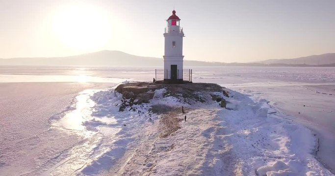 Flying above the frozen coasline, ascending aerial view of a lighthouse perched on a rocky Tokarevsky Spit. It's one of Vladivostok&rsquo;s main attractions. Russia. Winter. Aerial