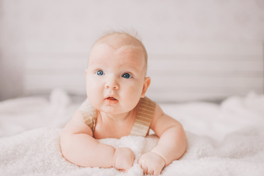 Adorable lovely newborn male baby boy with smiling emotional  happy face lifestyle indoor portrait. Funny infant child lying on stomach on bed with white wall on background.  Carefree childhood.