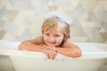 Little baby taking a bubble bath in a beautiful bathroom with a blue wall and flowers. Children's hygiene. Shampoo, hair treatment and soap for children. Baby bathes. The boy with foam in his hair.