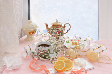 A cup of coffee with lemon and flowers in a festive ribbon, a plate with sweets on a snowy window,