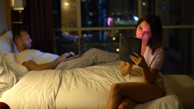 Couple in bed at night, woman using laptop, man sleeping, 4K
