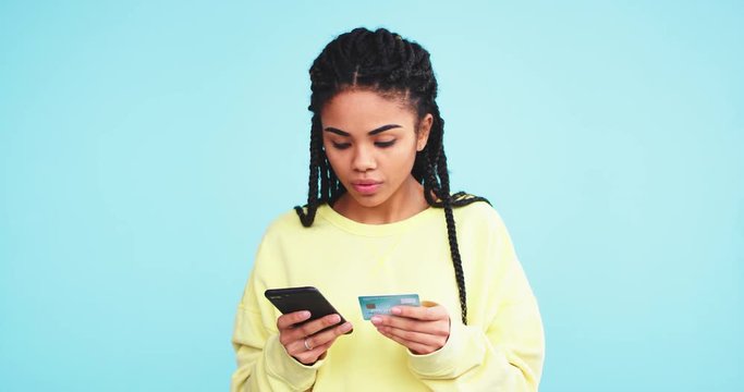 African pretty lady using a credit card and make a online order with her smartphone on a blue background wall.