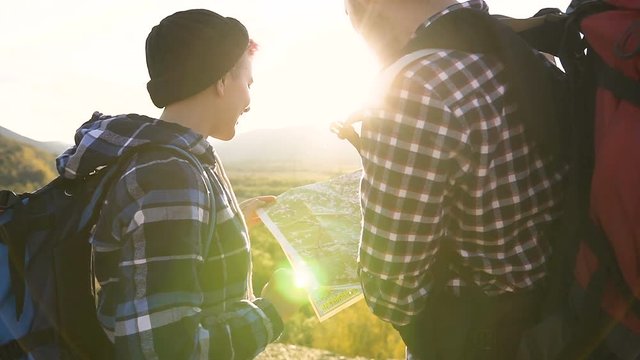 Back view. Close-up of young couple with backpacks and tourist map, standing in on top of mountain enjoying hiking trip to the mountains, trying to find direction