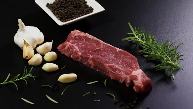 Meat Piece Garlic Rosmarinus And Pepper. A slow motion video of a raw steak, rosmarinus, garlic and pepper on a black table. The video moves slowly capturing the steak and seasonings.