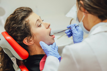 The doctor does professionally. Smiling beautiful girl with her white teeth, examined by a dentist in the clinic. A doctor is a good woman working in a mask and gloves.