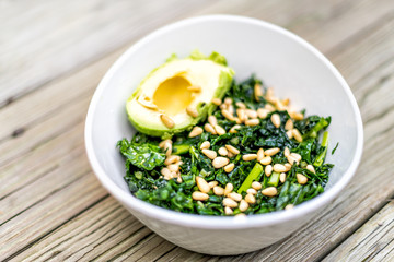 Closeup of kale green vegan vegetarian salad on wooden picnic table with pine nuts and avocado half in bowl