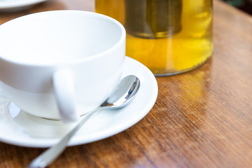 Closeup of one white cup on plates and green or oolong tea in breakfast brunch cafe restaurant wooden table with spoon and teapot or pot