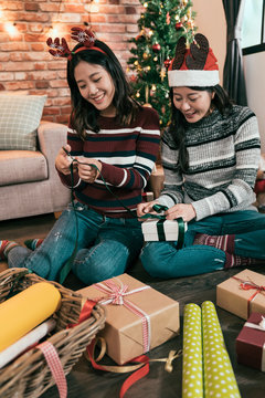 Female Friends Having Fun Doing Art Work Wrapping Gifts. Young Girl Using Scissors Cutting Green Ribbon Handmade Presents. Besties Prepare Celebration Christmas Eve New Year Beside Xmas Tree.