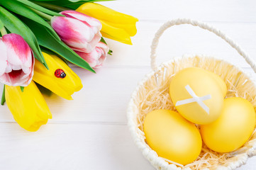 Colorful Easter eggs in white basket and bouquet of tulips. White wooden background. Easter holiday concept.