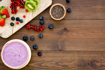 Cook trendy breakfast. Acai smoothie bowl near cutting board with fresh fruits, berries, chia seeds on dark wooden background top view copy space