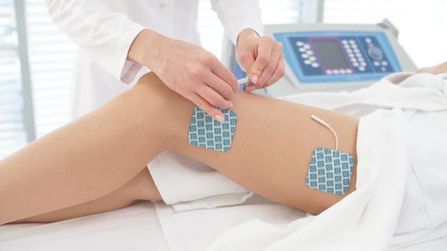 Beautician hands operating with Electric device, sticking electrodes to female hips. Myostimulation session performed on the slim female legs with healthy skin