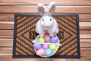 White bunny waiting on a doormat with a basket full of colorful Easter eggs
