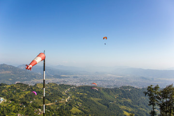 a red white windsock on the background of a flying paraglider in a clear blue sky and green...