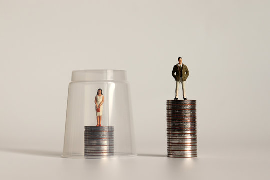 A glass ceiling concept. A miniature man and a miniature woman standing on a pile of coins of different heights.