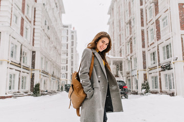 Stylish lady with brown backpack walking around city under snowfall. Outdoor photo of pretty girl with charming smile posing in gray coat on urban background in winter day.