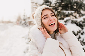True brightful emotions of winter girl in knittted hat smiling to camera on street full with snow. Closeup portrait, enjoying snow, waiting for christmas, cheerful mood, happiness