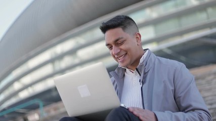 Smiling handsome young man using laptop. Happy middle eastern brunet with laptop on his knees sitting outdoor. Technology concept