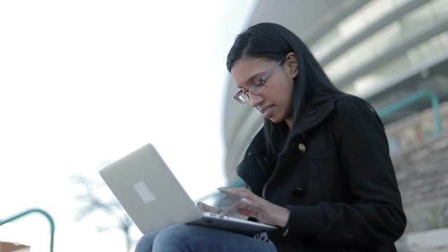 Smiling hindu woman in eyeglasses typing on laptop outdoor. Attractive brunette lady in eyeglasses working with laptop on her knees. Technology concept