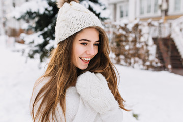 Cozy winter portrait of fashionable joyful young woman with long brunette hair walking on street full with snow. Surprised true positive emotions, warm white woolen gloves, knitted hat