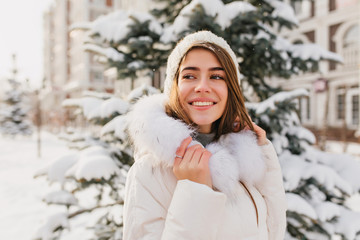 Inspired european lady wears white winter attire enjoying nature views. Outdoor portrait of stunning caucasian female model smiling on christmas tree background in weekend.