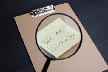 Closeup of magnifier and sticker "We`re hiring" on browm clipboard,black background.Concept of hiring new human to te vacant position