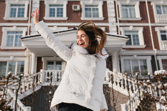 Inspired woman jumping in sunny winter day, enjoying christmas holidays. Outdoor photo of funny female model in white jacket chilling in weekend in frosty weather..